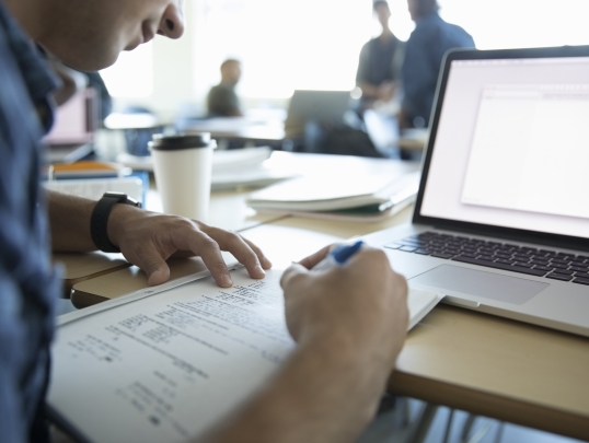 person writing at desk with an open laptop 